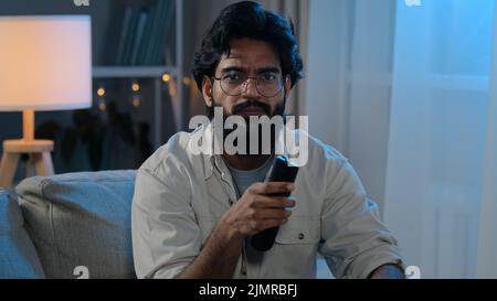 Evening entertainment Arabian Indian man bearded male in glasses in darkness living at home guy sits sofa switch change channel choosing program movie Stock Photo