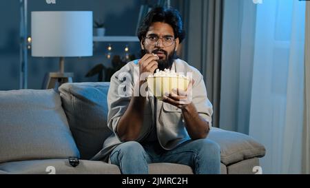 Serious interested Arabic Hispanic Indian bearded man guy wearing glasses 30s male with popcorn watching TV at home sofa late evening night relaxing Stock Photo