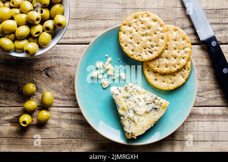 A slice of mature Stilton cheese on a plate with round whole wheat crackers and green olives in a small bowl to the side. A desert size serving Stock Photo