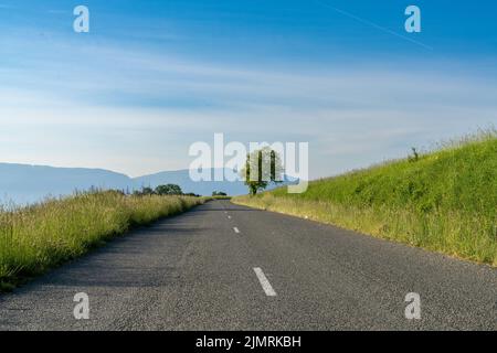 An empty blacktop highway in the countryside leading through green grass meadows to mountains in the background Stock Photo