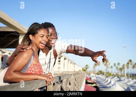 Beautiful couple of African origin have fun embracing in a coastal city center, leaning out of a gazebo pointing to the horizon. Background with copy Stock Photo