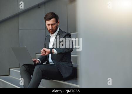 Focused businessman working laptop and looking on wriste watch sitting on stairs near office  Stock Photo