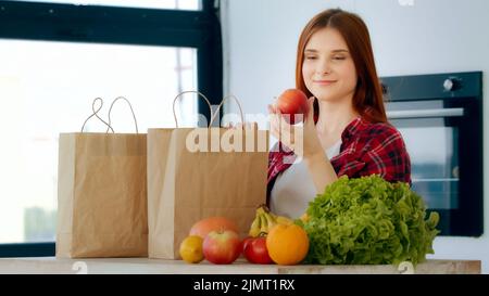 Female consumer woman caucasian buyer unpacks groceries from paper eco-packaging bag stands in modern kitchen. Cute redhead housewife take natural Stock Photo