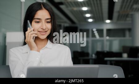 Busy female worker woman talk phone answer call arabic businesswoman do multitasking work on laptop chatting provide mobile support assistance for Stock Photo