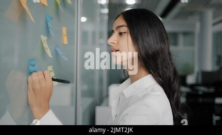 Thoughtful young businesswoman arabian hispanic creative designer come to note board in office read colorful stickers write reminders on sticky papers Stock Photo