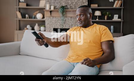 Elderly single bachelor african american adult senior man sitting on couch watching TV enjoy serial movie comments news program on television use Stock Photo