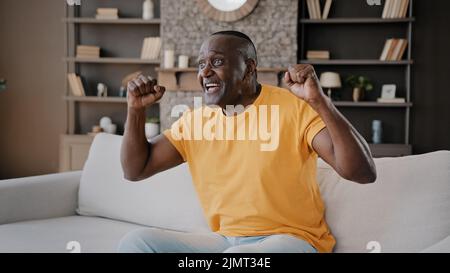 Senior football fan african american man bachelor sit on cozy sofa in apartment watch TV sport program hockey championship rooting for favorite team Stock Photo