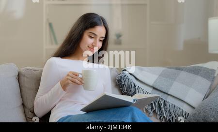 Serene young girl student daughter relaxed arabian biracial intelligent woman sits on sofa reads paper book scientific literature educational textbook Stock Photo
