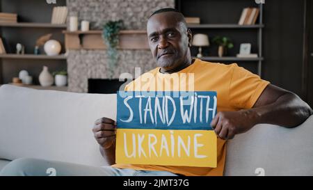 Serious elderly african american senior adult 50s man bachelor homeowner sits at home on sofa hold colorful cardboard in blue yellow colors painted Stock Photo