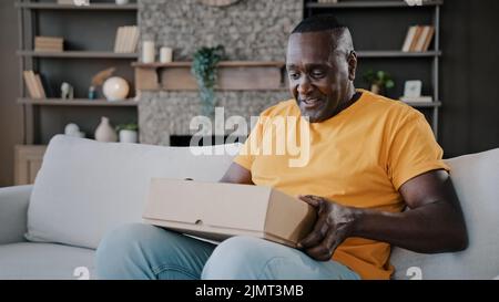 Surprised african adult man unpack parcel box american bachelor homeowner consumer birthday person male buyer get present from internet store delivery Stock Photo