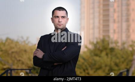 Portrait of confident strong caucasian businessman adult brunet man guy wears formal black suit posing with crossed arms outside handsome male Stock Photo