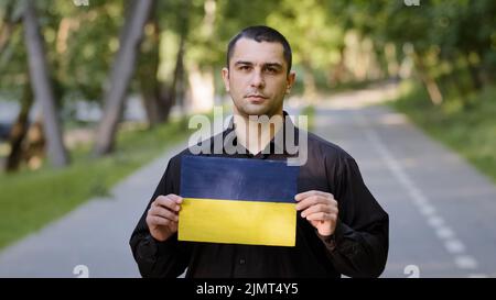 Serious man caucasian adult patriot activist protester wear black shirt stand outside in park hold cardboard drawing with blue yellow national flag of Stock Photo