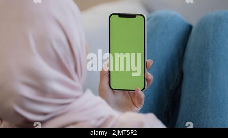 Rear view unknown islamic female muslim woman young lady girl in hijab lying on cozy sofa at home hold cellphone with green mobile screen display Stock Photo