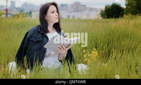 Intelligent caucasian woman sits on lawn outdoors reads book literature psychology bestseller educational encyclopedia textbook learns new language Stock Photo