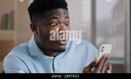 African american surprised man millennial 30s businessman employee owner hold mobile gadget modern device talk in video chat on smartphone shocked Stock Photo