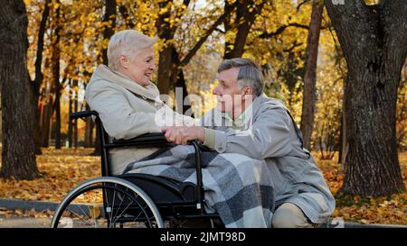 Caucasian senior man holding female hand caring woman with disability on wheelchair husband accompanies sick wife during rehabilitation in autumn park Stock Photo