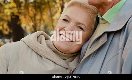 Happy middle aged pretty caucasian elderly woman grandma smiling stand relax in autumn park spend time outdoors lean on shoulder mature partner man Stock Photo