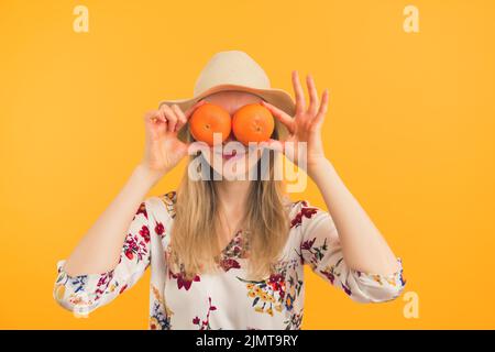 White woman with long blond hair wearing floral blouse and wide straw hat holding oranges in front of face. Studio shot. High quality photo Stock Photo