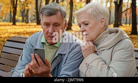 Upset old married caucasian couple use smartphone sit on bench in autumn park look at screen phone worried elderly retired people sad family feel shoc Stock Photo