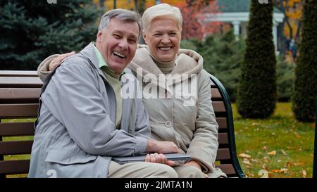 Caucasian married couple family retired 60s people man woman waving hands hello goodbye gesture elderly grandparents sit on bench in autumn park Stock Photo