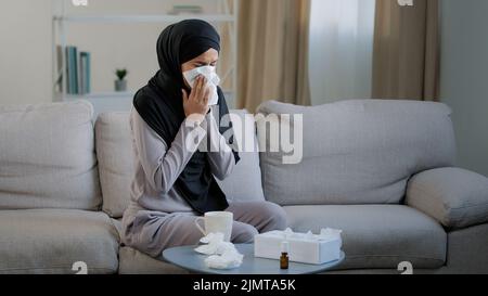 Sick arabian muslim young unhealthy woman in hijab suffer from runny nose flu disease seasonal allergy sneeze blow nose in paper napkin symptoms of Stock Photo