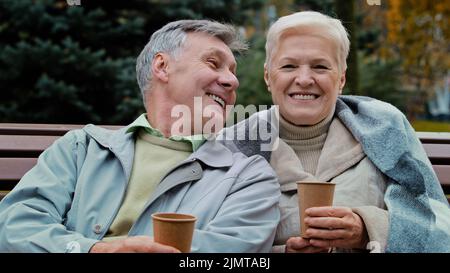 Happy senior aged couple talking sit in autumn park rest together carefree elderly grandparents drinking coffee tea outdoors enjoying pleasant convers Stock Photo