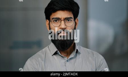 Portrait of serious handsome arabic hispanic young male bearded guy in eyeglasses wear shirt stands posing indoors looking at camera close-up unhappy Stock Photo