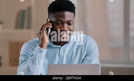 Smiling african man male businessman employee freelancer multitasking worker sit at home office talk on cellphone look at laptop screen provide remote Stock Photo