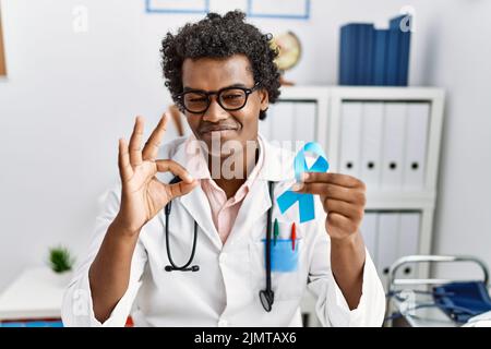 African doctor man holding blue ribbon doing ok sign with fingers, smiling friendly gesturing excellent symbol Stock Photo