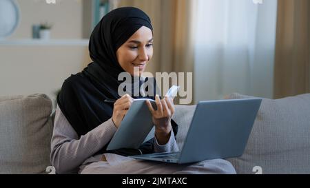 Cheerful young muslim arabian lady young woman sit on cozy sofa female freelancer writer take notes in diary noting information write book daily tasks Stock Photo