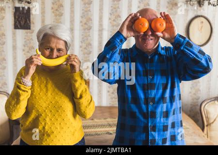 Funny caucasian senior couple - a wife holding a banana like a smile in front of her lips and her husband holding tangerines in front of his eyes. High quality photo Stock Photo