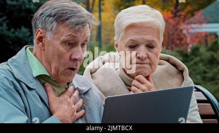 Shocked old married couple looking at laptop screen sitting in autumn park upset elderly man and woman reading bad news worried spouses aged feel shoc Stock Photo