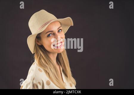 Young caucasian woman with long blond hair wearing a sun hat and linen shirt looking over the shoulder into camera with wide smile. Dark background studio shot. High quality photo Stock Photo