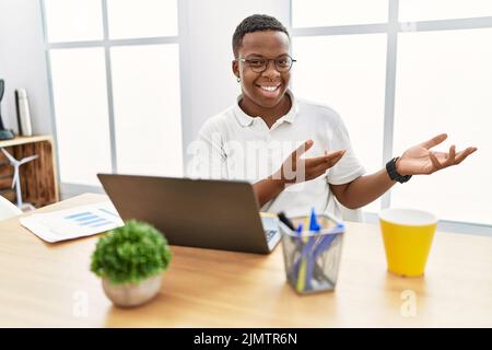 Young african man working at the office using computer laptop inviting to enter smiling natural with open hand Stock Photo