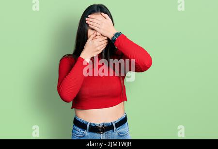 Young brunette teenager wearing red turtleneck sweater covering eyes and mouth with hands, surprised and shocked. hiding emotion Stock Photo