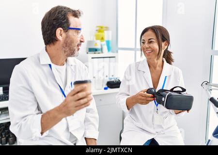 Middle age man and woman partners wearing scientist uniform drinking coffee and holding vr goggles at laboratory Stock Photo