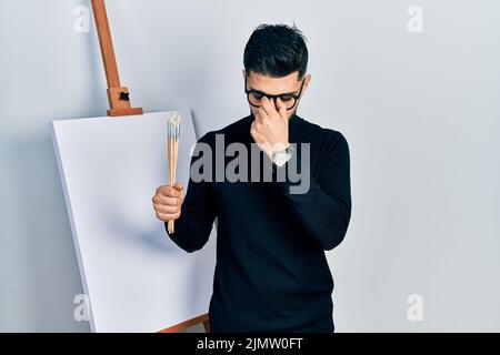 Handsome man with beard holding brushes close to easel stand tired rubbing nose and eyes feeling fatigue and headache. stress and frustration concept. Stock Photo