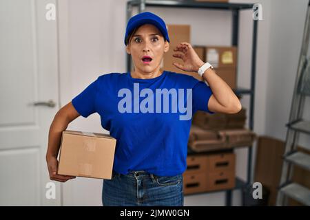Middle age brunette woman working wearing delivery uniform and cap crazy and scared with hands on head, afraid and surprised of shock with open mouth Stock Photo