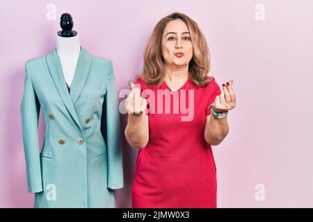 Middle age caucasian woman standing by manikin doing money gesture with hands, asking for salary payment, millionaire business Stock Photo