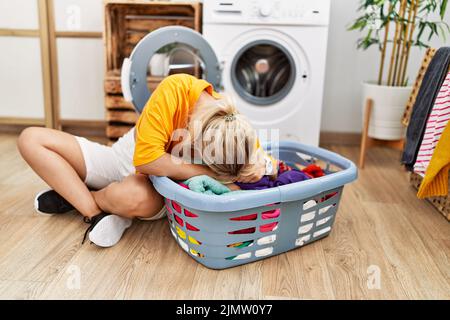 Young blonde girl doing laundry putting clothes into washing machine at home. Stock Photo