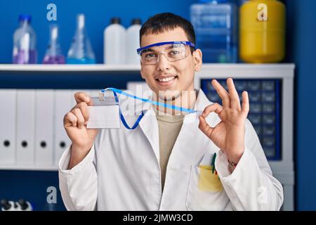 Young arab man working at scientist laboratory holding id card doing ok sign with fingers, smiling friendly gesturing excellent symbol Stock Photo
