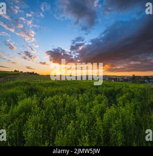 Summer countryside meadow, cloudy evening sunset sky, rural hills, village and fields in far. Stock Photo