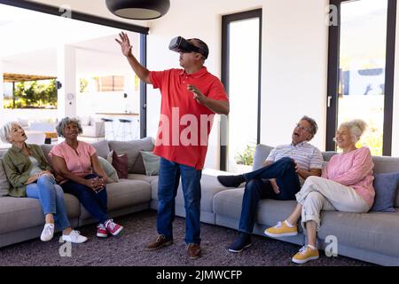 Multiracial friends looking at man using virtual reality simulator while sitting in retirement home Stock Photo