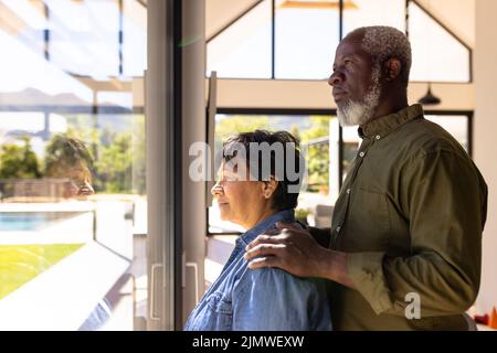 Side view of multiracial senior couple looking through window while standing in retirement home Stock Photo