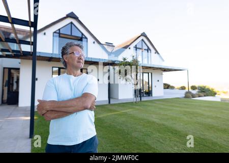 Thoughtful caucasian senior man with arms crossed standing against nursing home in yard, copy space Stock Photo