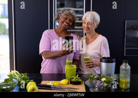 Smiling multiracial senior female friends looking at smartphone while standing by kitchen counter Stock Photo