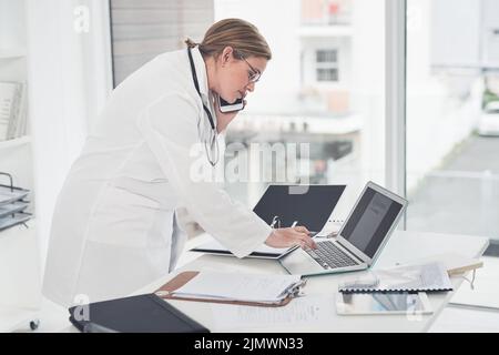 Let me just double-check that on my laptop. an attractive young female doctor typing on a laptop while taking a phonecall in her office. Stock Photo