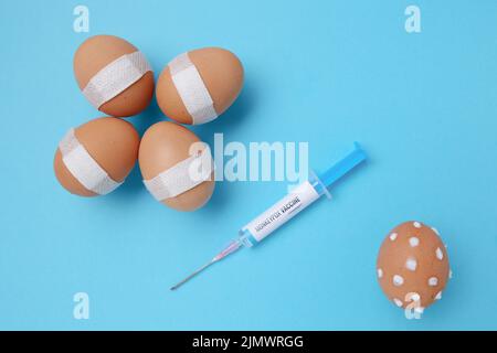 flatlay with monkeypox vaccine syringe, vaccinated eggs with medical patch and egg with monkeypox symptoms on blue background. Stock Photo
