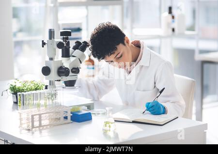 Recording of results is very important in science. an attractive young female scientist making notes while working with a microscope and plants in a Stock Photo