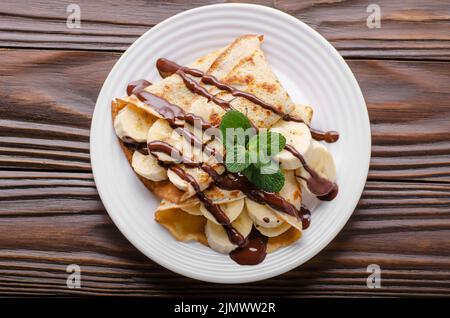 Flat lay French crepes with chocolate sauce and banana in ceramic dish on wooden kitchen table Stock Photo